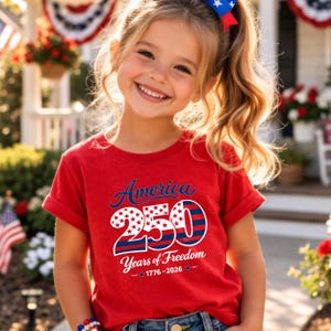 Może przedstawiać: Uśmiechnięta dziewczynka w czerwonej koszulce z napisem "America 250 Years of Freedom 1776-2026". Projekt zawiera gwiazdy i paski w kolorze niebieskim i białym. Ma blond włosy i kokardę w patriotycznych barwach.