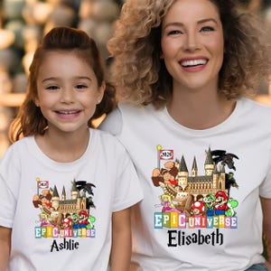 Puede incluir: Camisetas blancas con un gr&aacute;fico colorido que presenta un castillo, personajes de dibujos animados y el texto "EPIC UNIVERSE". Una camiseta tiene el nombre "Ashlie" y la otra "Elisabeth". Una peque&ntilde;a bandera muestra el a&ntilde;o "2026".