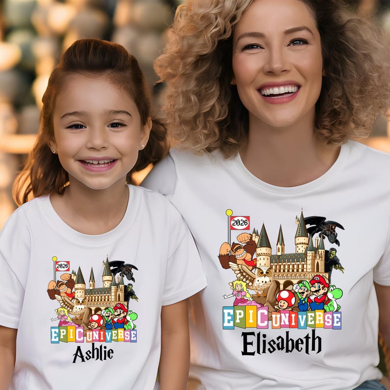 Puede incluir: Camisetas blancas con un gr&aacute;fico colorido que presenta un castillo, personajes de dibujos animados y el texto "EPIC UNIVERSE". Una camiseta tiene el nombre "Ashlie" y la otra "Elisabeth". Una peque&ntilde;a bandera muestra el a&ntilde;o "2026".