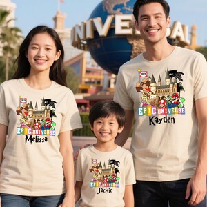 Puede incluir: Tres personas con camisetas beige a juego con un dise&ntilde;o gr&aacute;fico colorido. El dise&ntilde;o incluye un castillo, personajes de dibujos animados y el texto "EPIC UNIVERSE" con nombres individuales debajo. Un gran globo es visible en el fondo.