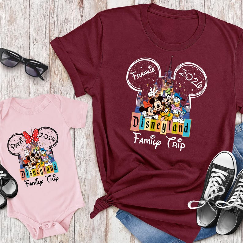 Puede incluir: Una camiseta granate y un body rosa con un dise&ntilde;o de "Disneyland Family Trip". El dise&ntilde;o incluye a Mickey y Minnie Mouse, el Pato Donald y Goofy. La camiseta tiene el nombre "Frankie" y el a&ntilde;o "2020" en un contorno de oreja de Mickey Mouse.