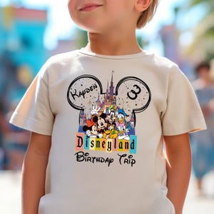Puede incluir: Camiseta beige de manga corta con un dise&ntilde;o en forma de Mickey Mouse, el nombre "Kayden" y el n&uacute;mero "3". El dise&ntilde;o incluye a Mickey, Minnie, Donald y Goofy frente a un castillo. Tambi&eacute;n est&aacute;n impresas las palabras "Disneyland Birthday Trip".
