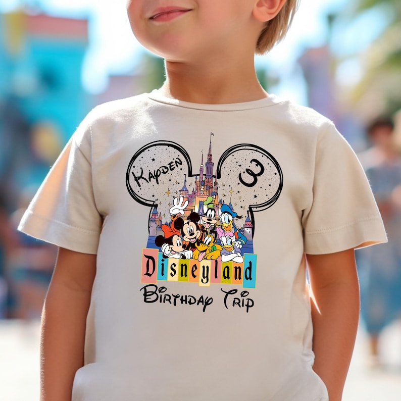 Puede incluir: Camiseta beige de manga corta con un dise&ntilde;o en forma de Mickey Mouse, el nombre "Kayden" y el n&uacute;mero "3". El dise&ntilde;o incluye a Mickey, Minnie, Donald y Goofy frente a un castillo. Tambi&eacute;n est&aacute;n impresas las palabras "Disneyland Birthday Trip".