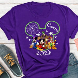 May include: A purple short-sleeved t-shirt featuring a graphic of Disney characters in Halloween costumes inside a lime green Mickey Mouse ear outline. The design includes the name Carlos and the year 2026 in white text with a spider web.