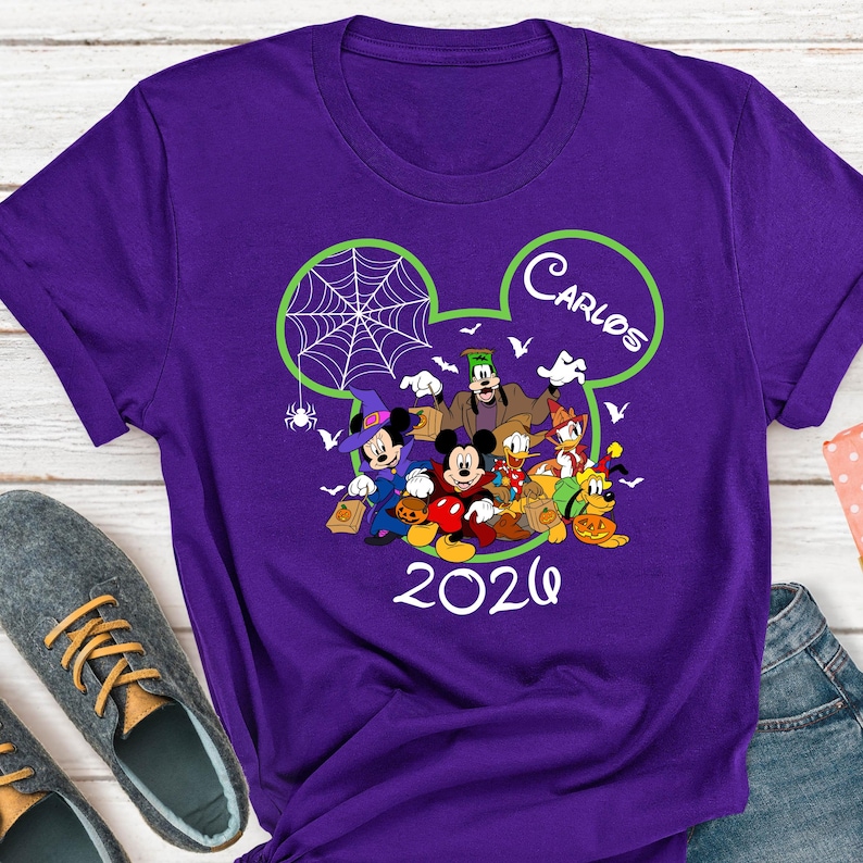 May include: A purple short-sleeved t-shirt featuring a graphic of Disney characters in Halloween costumes inside a lime green Mickey Mouse ear outline. The design includes the name Carlos and the year 2026 in white text with a spider web.
