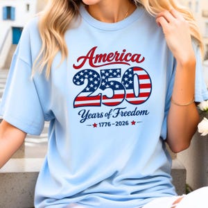 Może przedstawiać: Jasnoniebieska koszulka z napisem "America 250 Years of Freedom 1776-2026" w kolorze czerwonym, białym i niebieskim. Liczba "250" jest zaprojektowana z gwiazdami i paskami.