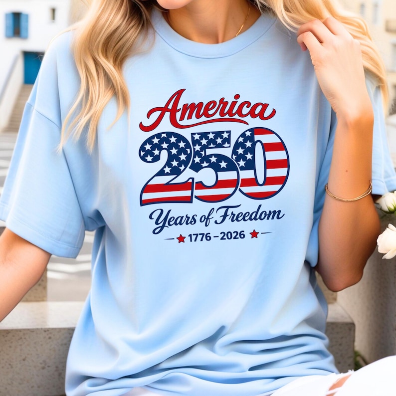 Może przedstawiać: Jasnoniebieska koszulka z napisem "America 250 Years of Freedom 1776-2026" w kolorze czerwonym, białym i niebieskim. Liczba "250" jest zaprojektowana z gwiazdami i paskami.