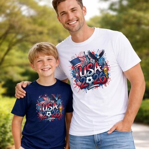 May include: Two people wearing matching t-shirts with a patriotic design. The shirts feature the text "USA 2026" with a soccer ball, American flag, eagle, and other patriotic elements. One shirt is white, the other is navy blue.
