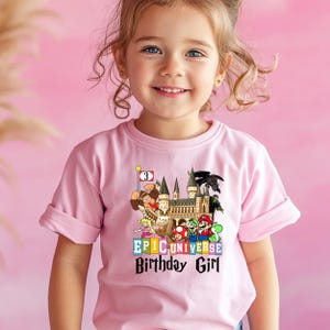 Puede incluir: Camiseta rosa claro con un gr&aacute;fico colorido. El gr&aacute;fico incluye personajes de dibujos animados, un castillo y el texto "EPIC UNIVERSE Birthday Girl". Una bandera muestra el n&uacute;mero "3".