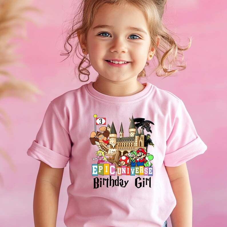 Puede incluir: Camiseta rosa claro con un gr&aacute;fico colorido. El gr&aacute;fico incluye personajes de dibujos animados, un castillo y el texto "EPIC UNIVERSE Birthday Girl". Una bandera muestra el n&uacute;mero "3".