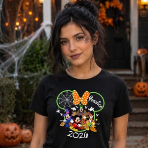 May include: A black t-shirt featuring a graphic of Mickey Mouse and friends in Halloween costumes inside a green Mickey-shaped outline. The design includes a spiderweb, an orange polka-dot bow, the name Jessica, and the year 2026 in white text.