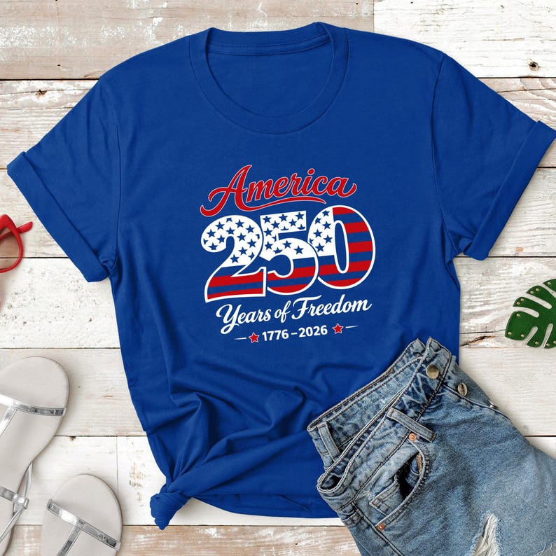 Może przedstawiać: Kr&oacute;lewski niebieski t-shirt z napisem "America 250 Years of Freedom 1776-2026" w kolorze czerwonym, białym i niebieskim. Projekt zawiera słowo "America" w skrypcie nad liczbą "250" z gwiazdami i paskami.