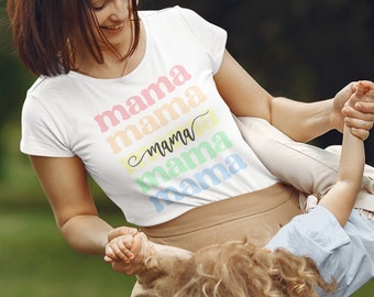 Puede incluir: Camiseta blanca con la palabra "mama" en colores arcoíris. La palabra "mama" se repite varias veces en un diseño apilado. La persona lleva la camiseta y sostiene a un niño.