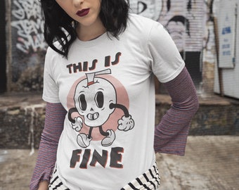 May include: A white t-shirt with a cartoon skull graphic and the text "This is Fine". The shirt is tied at the waist with a black and white striped belt. The person wearing the shirt has black hair and is standing in front of a graffiti wall.