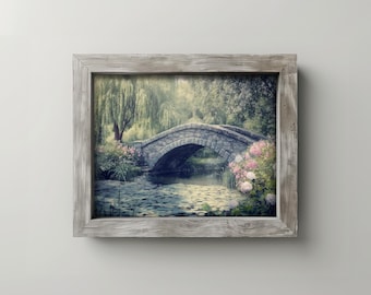 May include: A framed photograph of a stone bridge over a pond with lily pads. The bridge is surrounded by lush greenery and pink flowers.