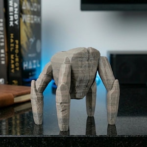 May include: A gray, 3D-printed sculpture of a four-legged creature with a textured, rock-like appearance. The sculpture has a blocky body and articulated limbs, resembling a stylized robot or alien figure. The sculpture is displayed on a dark surface.