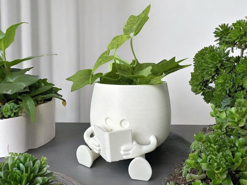 May include: White ceramic planter shaped like a person sitting with legs crossed and holding a book. The planter has two small eyes and is holding a white book. The planter is filled with green leafy plants.
