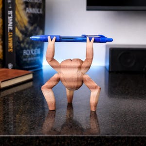 May include: A whimsical wooden pen holder shaped like a figure lifting a blue pen overhead. The pen holder is light brown and has a unique, artistic design. The pen is a standard blue ballpoint pen.
