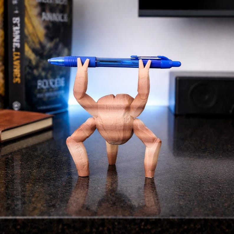 May include: A whimsical wooden pen holder shaped like a figure lifting a blue pen overhead. The pen holder is light brown and has a unique, artistic design. The pen is a standard blue ballpoint pen.