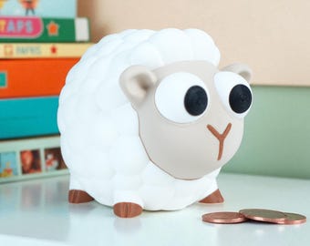May include: A white sheep-shaped piggy bank with a light brown face, ears, and hooves. The sheep has large black eyes and a Y-shaped nose. Several coins are in front of the bank. The bank is on a white surface.
