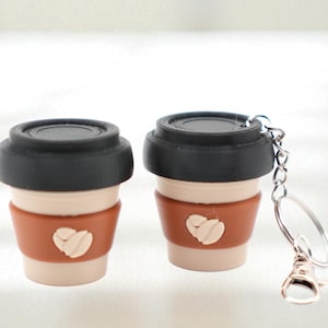 May include: Two coffee cup keychains. The cups have a black lid, a tan body, and a brown sleeve with a white heart. One keychain has a silver chain and clasp.
