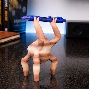 May include: A whimsical, wooden sculpture of a figure with arms and legs, holding a blue pen. The figure is brown with visible wood grain. The pen is a vibrant blue with silver accents and a clear tip. The sculpture is on a dark surface.