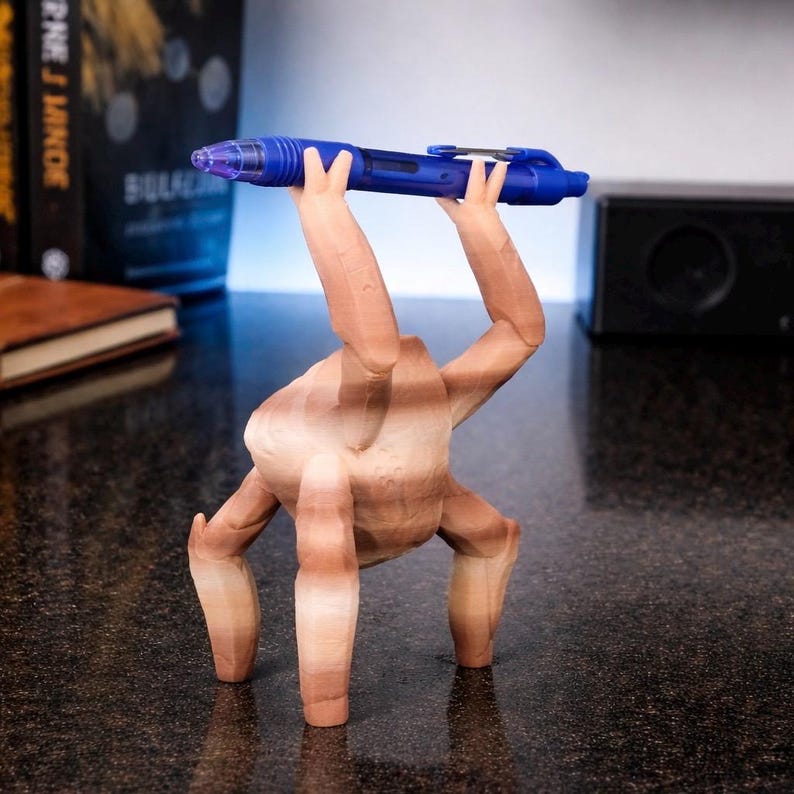 May include: A whimsical, wooden sculpture of a figure with arms and legs, holding a blue pen. The figure is brown with visible wood grain. The pen is a vibrant blue with silver accents and a clear tip. The sculpture is on a dark surface.