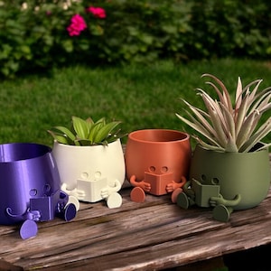 May include: Four colorful ceramic planters shaped like people sitting with their legs crossed and reading a book. The planters are purple, white, orange, and green.