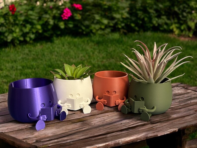 May include: Four colorful ceramic planters shaped like people sitting with their legs crossed and reading a book. The planters are purple, white, orange, and green.