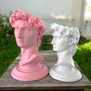 May include: Two plaster sculptures of the David statue, one in pink and one in white, with a glittery finish. The sculptures are on a wooden surface.