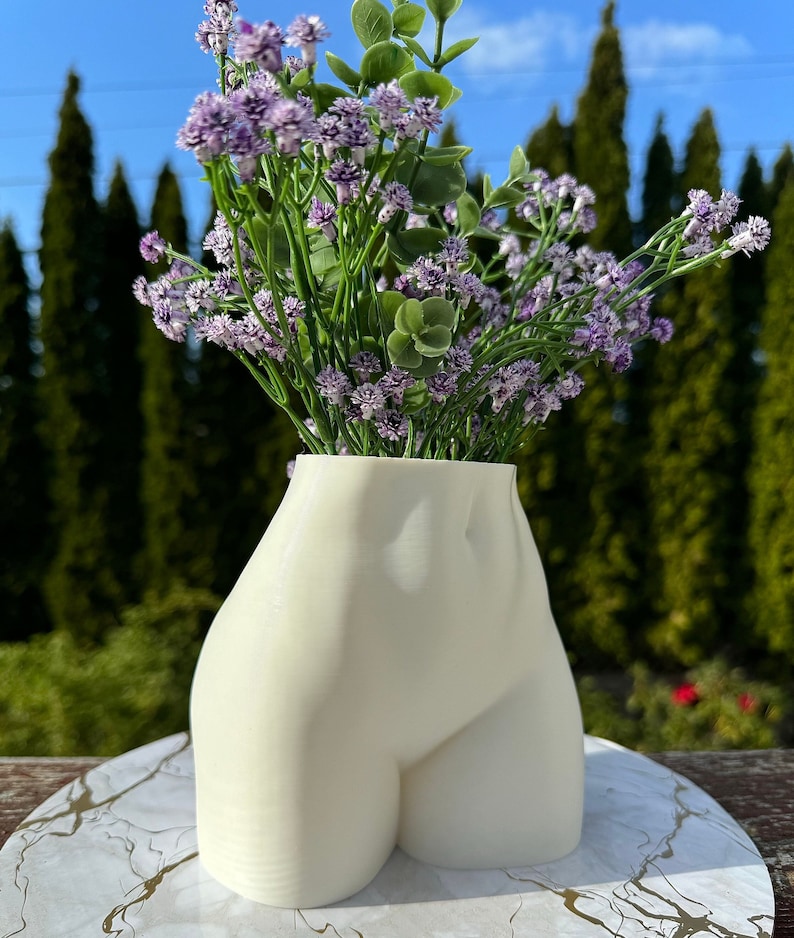 May include: White ceramic vase shaped like a human torso, with a bouquet of purple flowers.