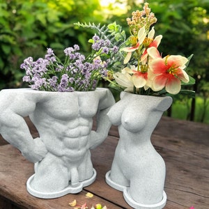 Könnte beinhalten: Zwei graue Beton-Blumenkästen in Form von menschlichen Torsos. Der Blumenkasten links ist ein männlicher Torso mit definierten Muskeln. Der Blumenkasten rechts ist ein weiblicher Torso. Beide Blumenkästen haben Blumen, die aus ihnen herauswachsen.