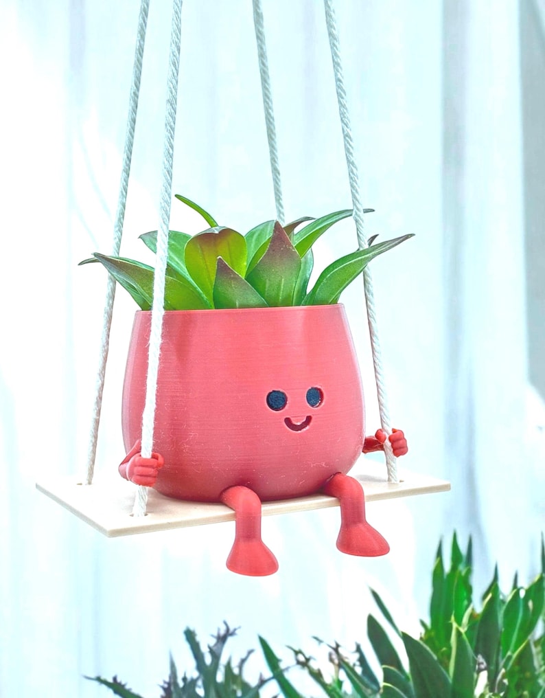 May include: A pink, smiling planter with legs and arms, holding onto a white swing. The planter is filled with a green succulent plant.