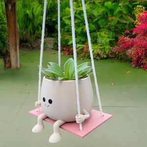 May include: A white ceramic planter with a smiling face and legs, hanging from a white rope and holding a green succulent plant. The planter is sitting on a pink wooden swing.