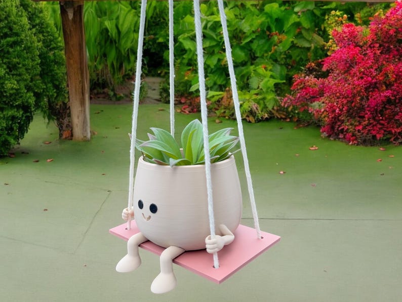 May include: A white ceramic planter with a smiling face and legs, hanging from a white rope and holding a green succulent plant. The planter is sitting on a pink wooden swing.