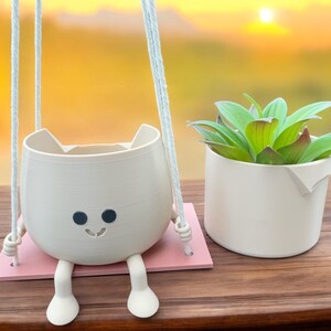 May include: A white ceramic planter shaped like a cat with a smiling face and black eyes, hanging from a white rope. The planter is on a pink platform and has two white legs. A green succulent plant is in a white ceramic pot next to the hanging planter.