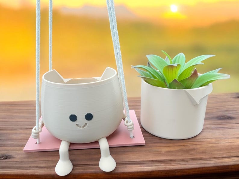 May include: A white ceramic planter shaped like a cat with a smiling face and black eyes, hanging from a white rope. The planter is on a pink platform and has two white legs. A green succulent plant is in a white ceramic pot next to the hanging planter.