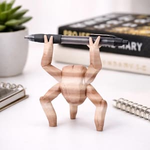 May include: A wooden pen holder shaped like a stylized figure with four legs and arms raised to hold a black pen. The pen has a black grip and a clear barrel. The holder is light brown with wood grain.