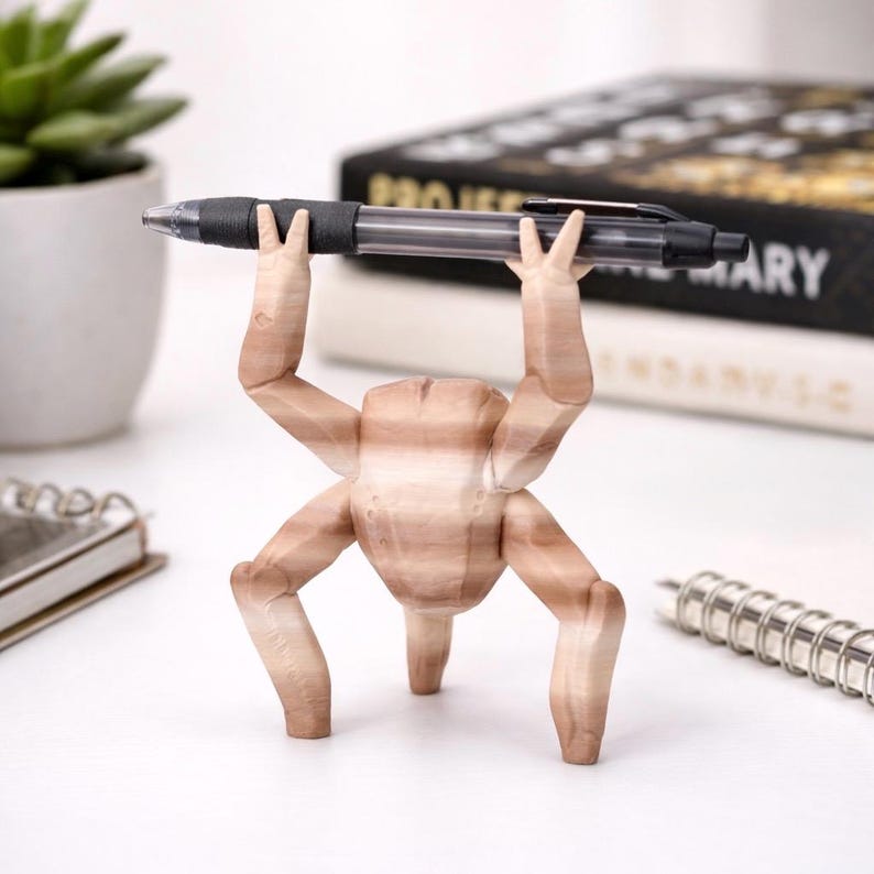 May include: A wooden pen holder shaped like a stylized figure with four legs and arms raised to hold a black pen. The pen has a black grip and a clear barrel. The holder is light brown with wood grain.