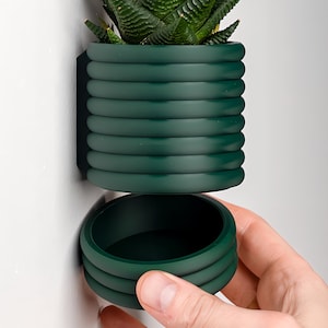 May include: A dark green, cylindrical planter with a removable bottom section. The planter is made of a smooth, textured material and has a modern, minimalist design.