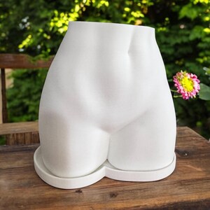 May include: White ceramic planter shaped like a human torso. The planter has a smooth, rounded surface and a wide opening at the top for planting.