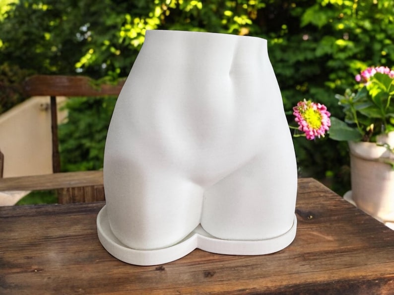 May include: White ceramic planter shaped like a human torso. The planter has a smooth, rounded surface and a wide opening at the top for planting.