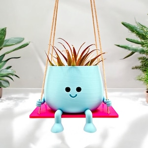 May include: A light blue planter shaped like a smiling face with legs, hanging from a rope swing. The planter is holding a green plant with brown leaves. The planter is sitting on a pink platform.
