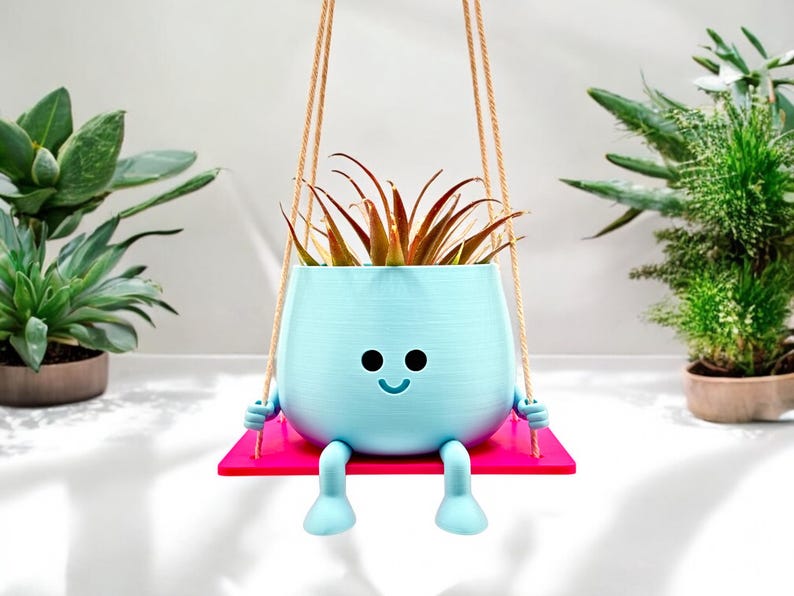 May include: A light blue planter shaped like a smiling face with legs, hanging from a rope swing. The planter is holding a green plant with brown leaves. The planter is sitting on a pink platform.