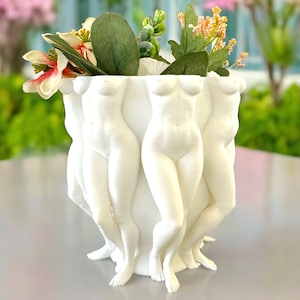 Può includere: Vaso in ceramica bianca a forma di tre figure femminili in piedi schiena contro schiena. Il vaso è pieno di fiori artificiali.