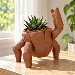 May include: A brown, rock-textured planter shaped like a creature with articulated limbs and a raised claw, holding a small green succulent plant with white horizontal stripes on its pointed, fleshy leaves.