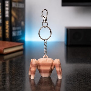 May include: A light brown keychain featuring a stylized, geometric figure with four legs. The figure is attached to a silver keyring and clasp. The keychain is made of a light brown material and is displayed against a dark surface.
