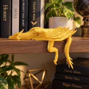 May include: A bright yellow dragon figurine with textured scales, folded wings, and a long tail, positioned to hang over the edge of a wooden shelf. The dragon has a detailed head with small horns and is crafted in a solid, matte finish.