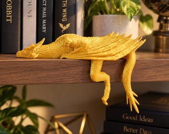 May include: A bright yellow dragon figurine with textured scales, folded wings, and a long tail, positioned to hang over the edge of a wooden shelf. The dragon has a detailed head with small horns and is crafted in a solid, matte finish.