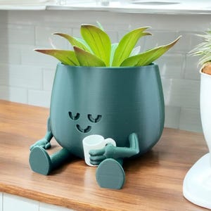 Op de afbeelding: Een donkergroene keramische plantenpot in de vorm van een persoon met een glimlachend gezicht en gesloten ogen. De pot houdt een witte keramische mok vast in de ene hand en heeft een kleine vetplant die erin groeit. De pot staat op een houten oppervlak.